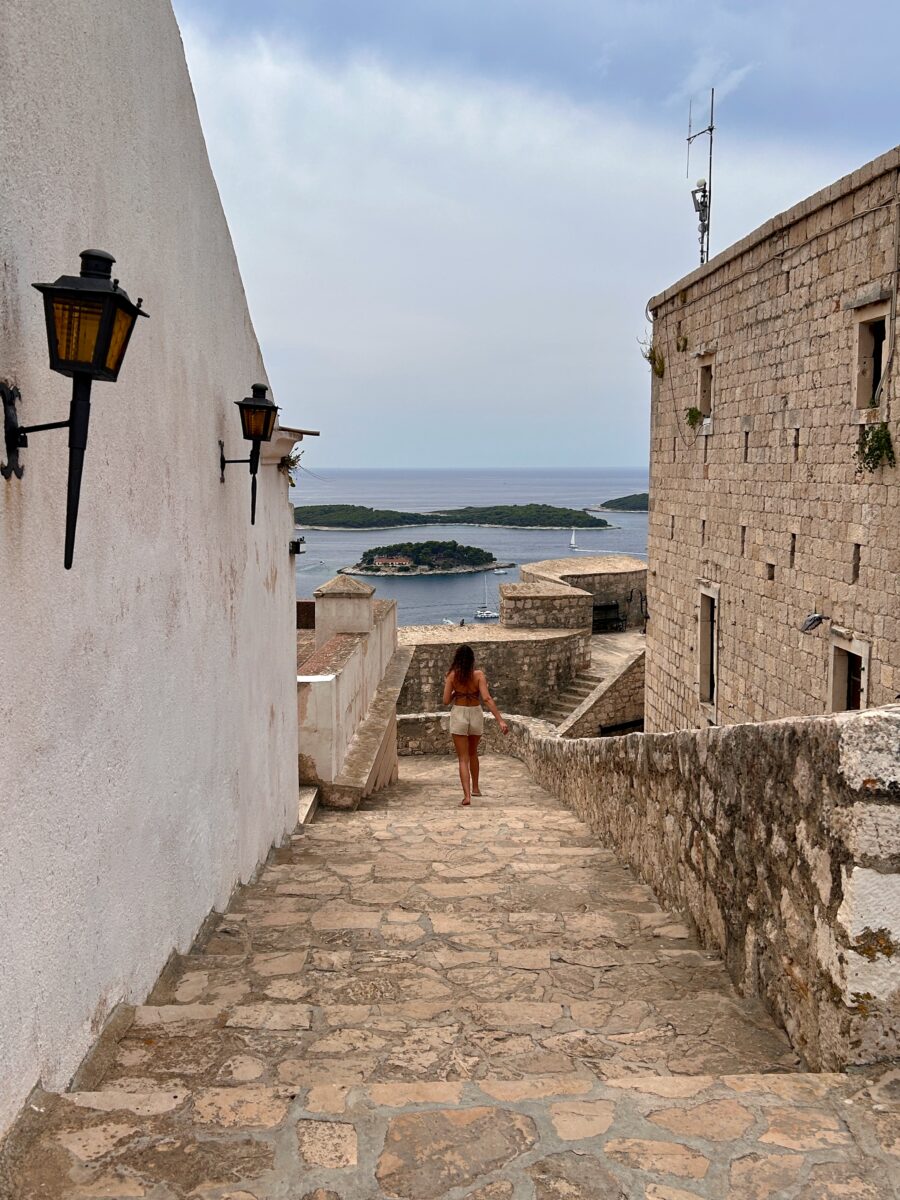 a woman walking down the stairs of the castle of Hvar, overlooking the marina and its nearby islands