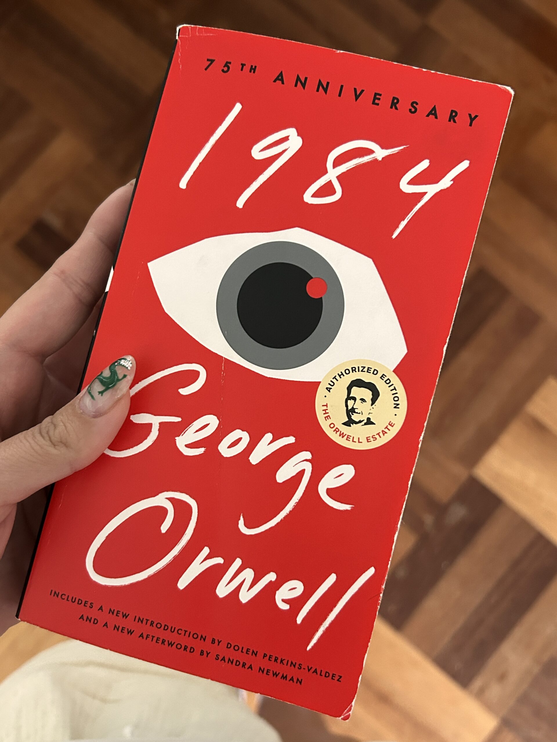 a feminine hand with nail art holding the book "1984" by George Orwell. The book cover is red and features a big eye.