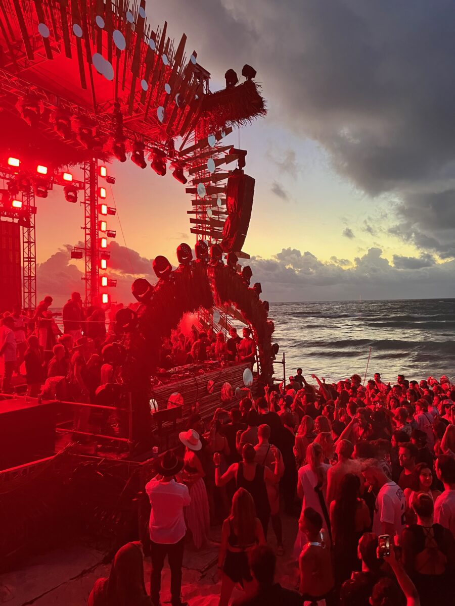 a crowd in front of a DJ stage on the beach of tulum at sunrise