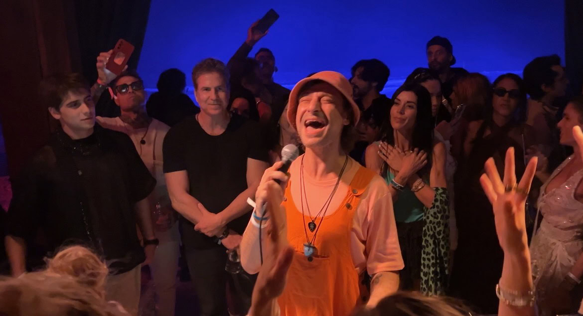 a man wearing orange overalls singing in front of a DJ booth in Tulum