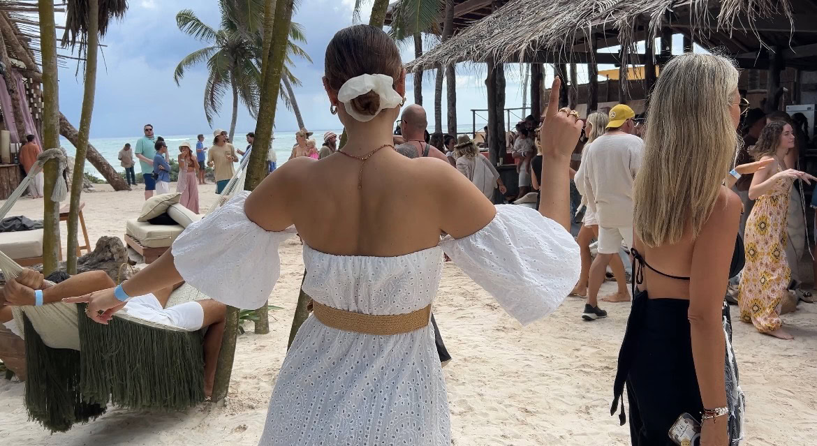 view of the back of a woman with a white dress dancing on the beach of tulum surrounded by people and palm trees